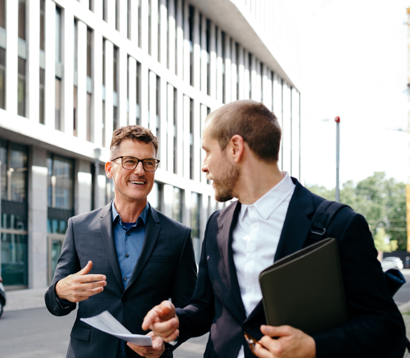 Two businessmen in suits walk and talk on a city street, holding documents and a laptop. They appear engaged and professional, with a modern building in the background.