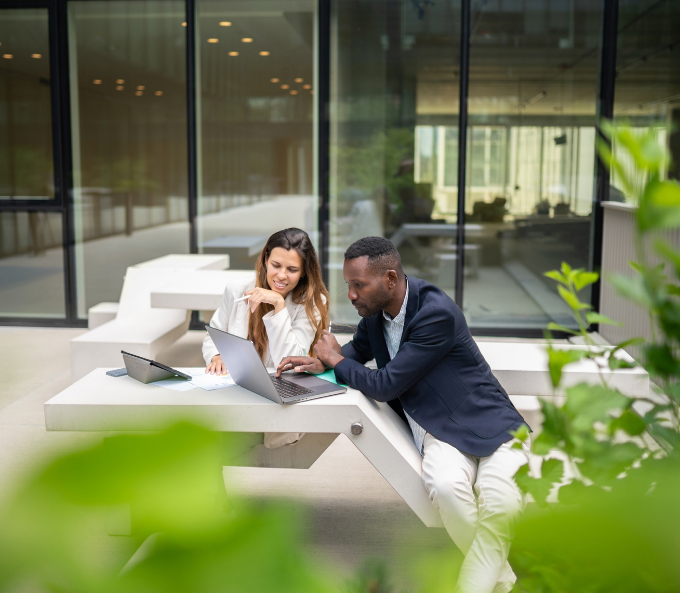 A man and woman sit at an outdoor table, focused on a laptop. They are surrounded by greenery, with large windows in the background and a collaborative expression.