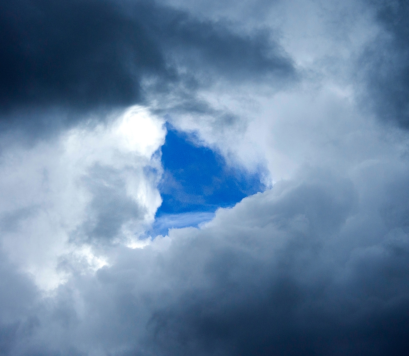 A circle of dark, stormy clouds frames a patch of vibrant blue sky, creating a dramatic contrast and conveying a sense of hope amid turbulence.