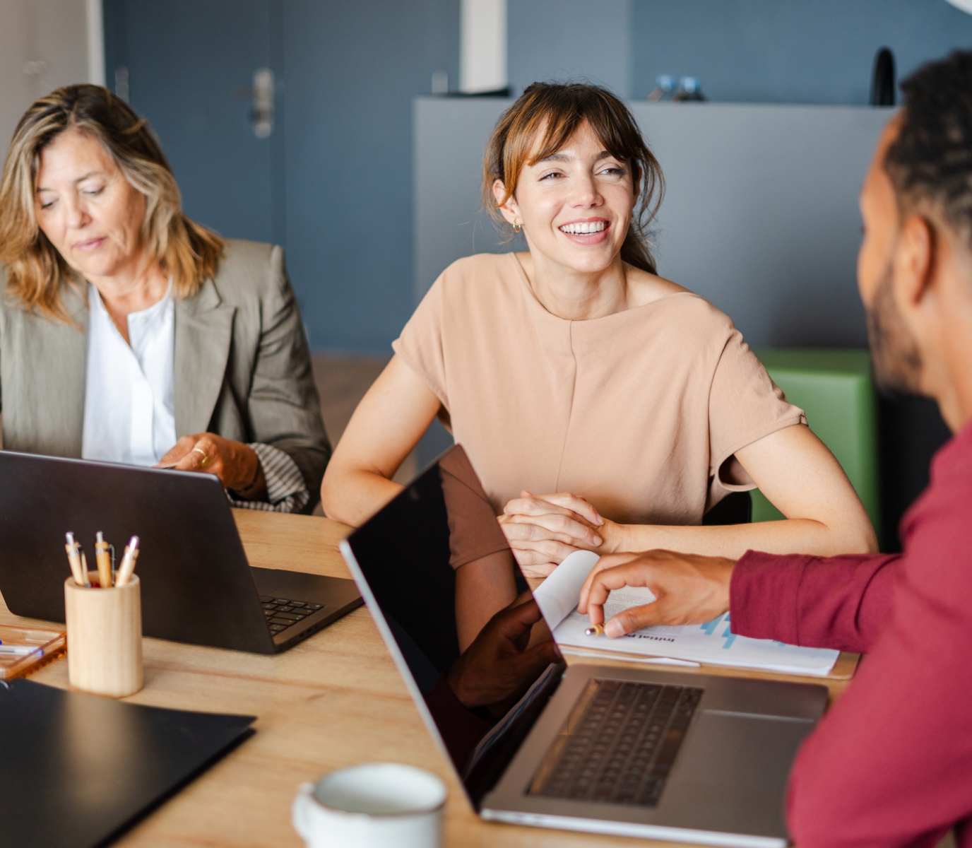Three colleagues are engaged in a meeting around a table with laptops and papers. The atmosphere is positive, with one person smiling warmly.