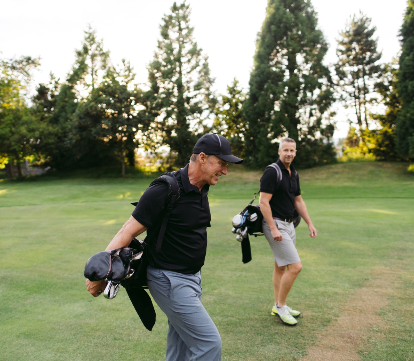 Two men walk on a golf course, smiling and carrying golf bags. They wear black shirts and caps, surrounded by lush green trees, under a bright sky.
