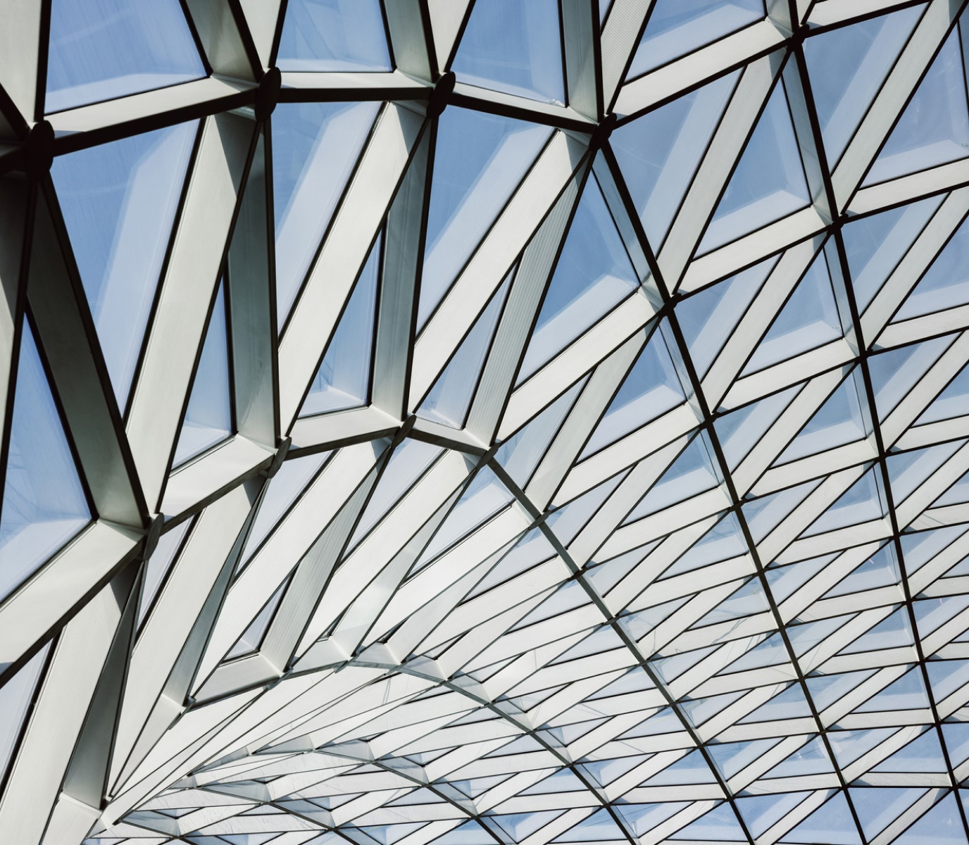 Geometric glass ceiling with triangular patterns against a blue sky, creating a modern and airy architectural feel. Light shines through, enhancing a sense of openness.