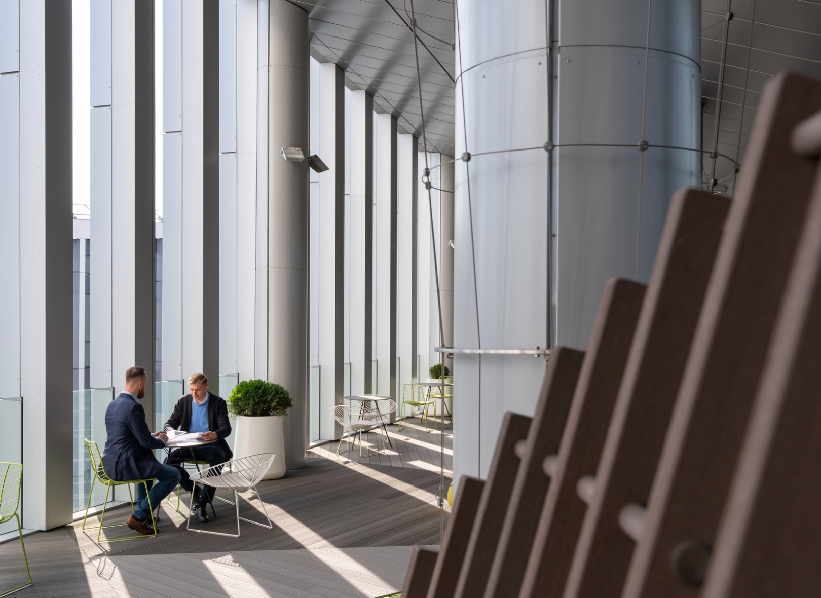 Two men in suits converse at a table in a modern, sunlit atrium with tall glass and metal walls, creating a serene and professional environment.